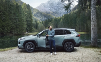 Mężczyzna wysiada z Toyoty nad jeziorem w lesie i podziwia widok, w tle góry